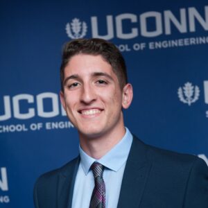 Rising senior Steven Porter stands in a suit jacket and tie in front of a blue background on which the UConn wordmark printed.