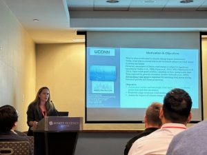 Marina Astitha delivers a presentation at a professional conference, speaking at a podium with a projected research slide titled “Motivation & Objectives” in the background. Audience members are seated and listening.