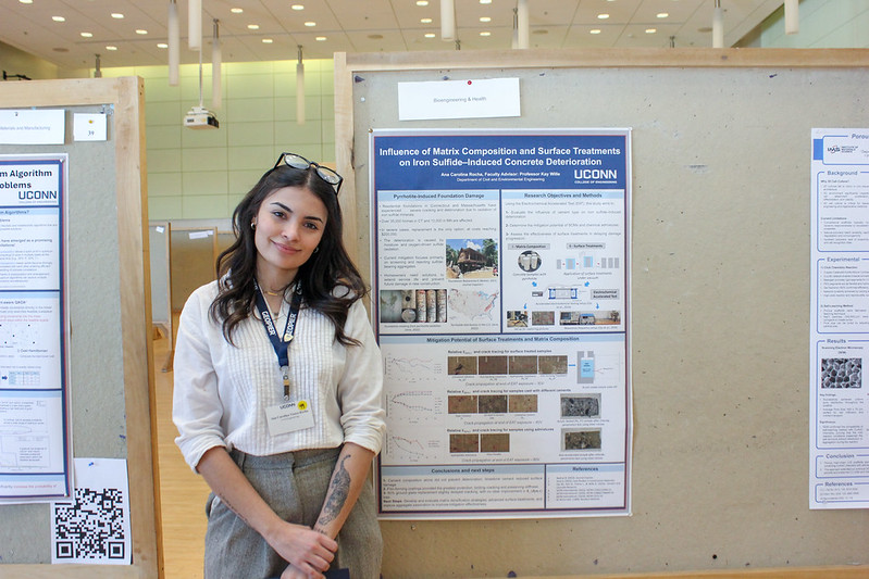 A student stands in front of her research poster, smiling at the camera in a poster session setting, with other displays visible nearby.