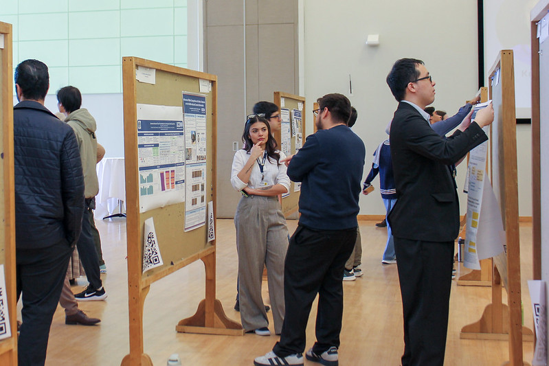 Several students stand beside large poster boards arranged in rows, discussing research and reviewing posters in an indoor exhibition space.