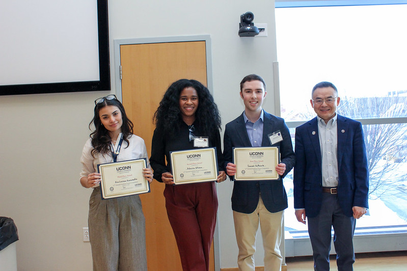 Three award-winning students stand side by side holding certificates next to a faculty member, posing for a photo in front of a large window.