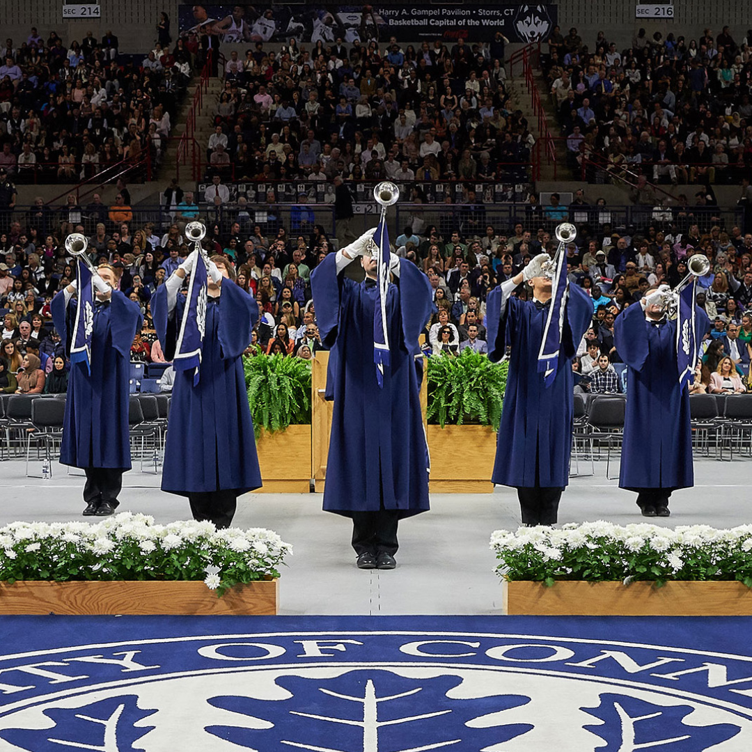 Five ceremonial trumpeters in blue robes stand in a line at the front of a graduation ceremony, playing silver trumpets. They are on a stage with white flowers and plants, with a large audience seated behind them in an arena.