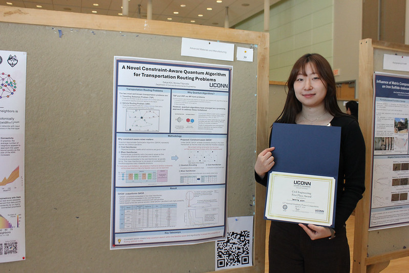 A student stands beside her research poster, holding a certificate and smiling, with other posters visible in the background.