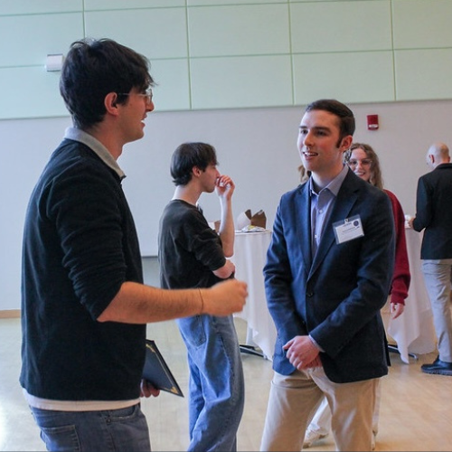 Two students stand in a bright indoor space during a poster session, engaged in conversation; one wears a blazer and name badge, while the other holds a tablet, with additional attendees and posters visible in the background.