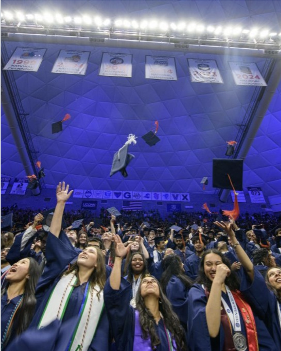 A large group of graduates in navy blue caps and gowns celebrate inside a domed arena, tossing their mortarboards into the air. Many are smiling, cheering, and reaching upward as the caps scatter above them, some with orange tassels visible. The crowd fills the foreground, while bright stadium lights and championship banners hang overhead in the background, emphasizing the scale and excitement of the commencement ceremony.