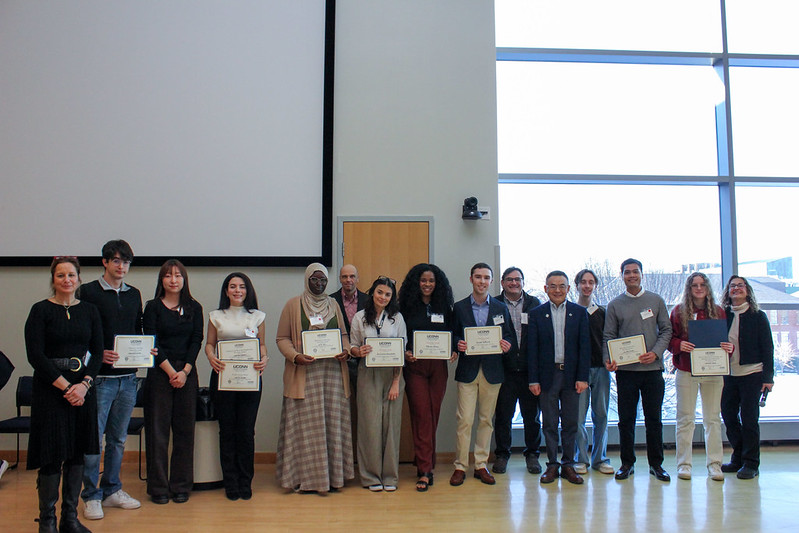 A group of graduate students and faculty stand in a row at the front of a large room, several holding certificates, posing for a photo after the poster competition awards.