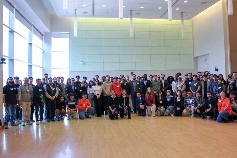 A large group of students, faculty, and staff gather in a spacious, well-lit hall for a group photo, representing participants in the poster competition.