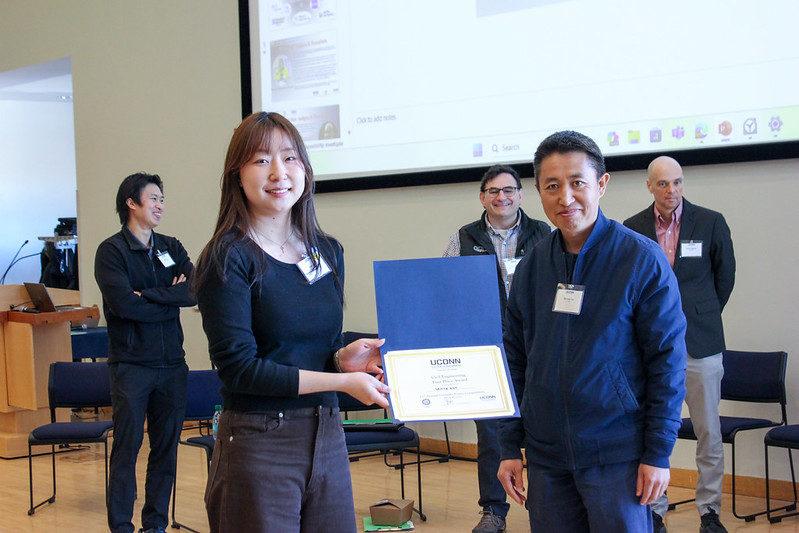 A student holds a certificate while standing next to a faculty member during an award presentation, with several people and a projection screen visible in the background.