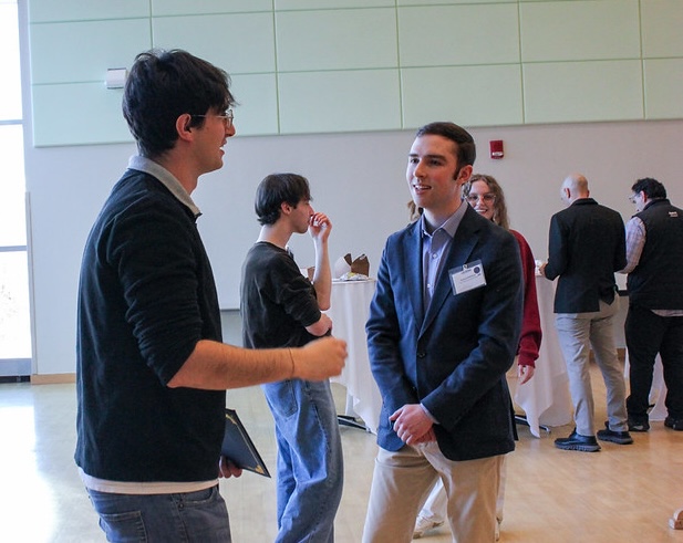 Two students stand in a bright indoor space during a poster session, engaged in conversation; one wears a blazer and name badge, while the other holds a tablet, with additional attendees and posters visible in the background.