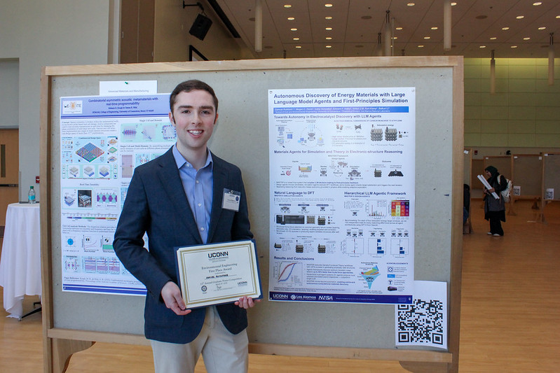 A graduate student stands smiling in front of a research poster display, holding a framed certificate, with the poster mounted on a wooden board behind him.